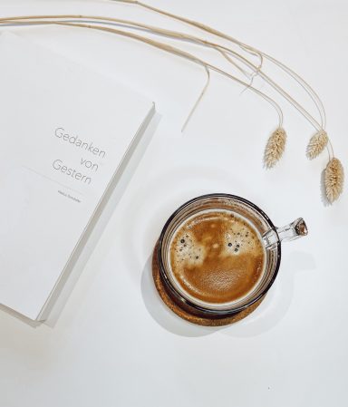 Ein Becher Kaffee steht neben einem offenen Buch und dekorativen Gräsern.